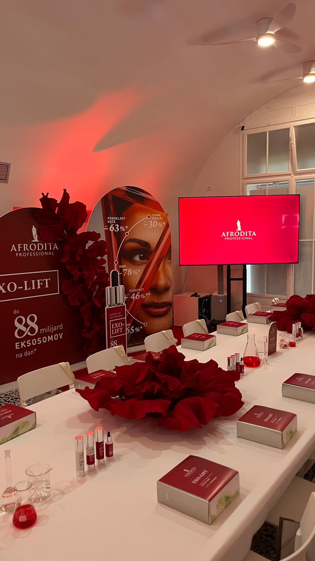 Afrodita Professional EXO-LIFT lansiranje v 369 Place Ljubljana — dolga miza z brandingom, rdeči cvetlični aranžma, vaultani strop